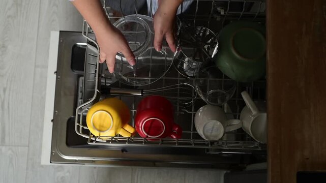 Top view of woman opening dishwasher, pulling out upper rack and removing clean cooking utensils. Overhead kitchen routine showing unloading of heat resistant tools at home.
