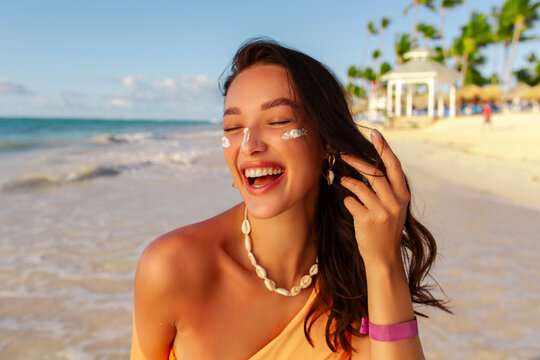 Skin care, sun protection concept. Joyful woman with sun cream on face, standing on sandy beach with turquoise water and palms on background