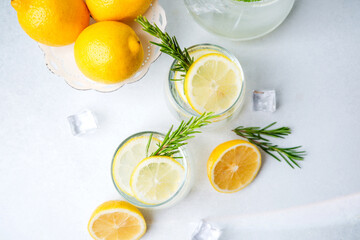 Refreshing lemon water in clear glasses with rosemary sprigs ice cubes and fresh lemon halves on white background