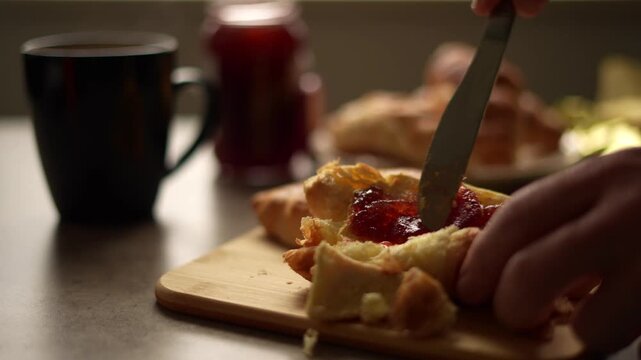 Spreading jam on buttered croissants on a cutting board with cup of hot tea.