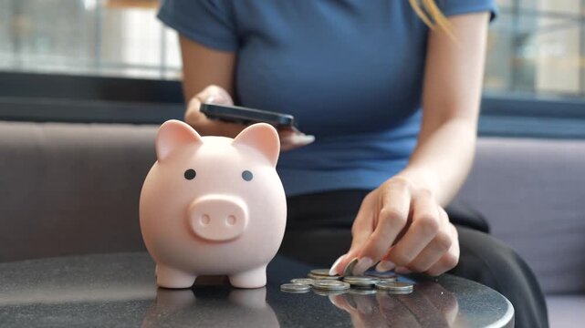 A woman puts a coin into a piggy bank beside her smartphone, representing saving money, digital finance, and smart personal budgeting.