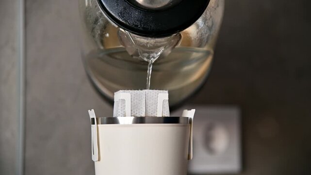 Water being poured from a glass kettle into a white filter holder containing filter papers, positioned on a countertop with a modern kitchen backdrop