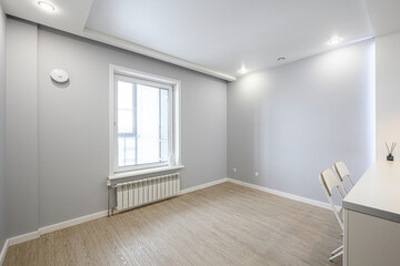 Fototapeta premium A minimalist room with light grey walls, wooden flooring, large window with white frame, radiator, recessed ceiling lights, white desk and chairs, smoke detector