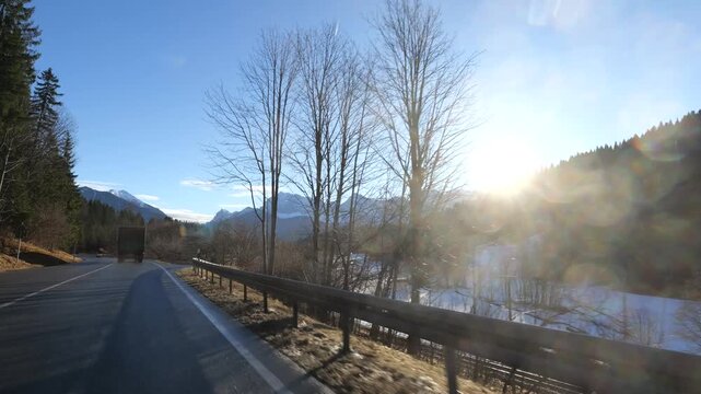truck travels winding alpine road seefeld framed by bare winter trees brilliant sunburst reflecting off snow dusted valley distant jagged peaks down tirol austria 