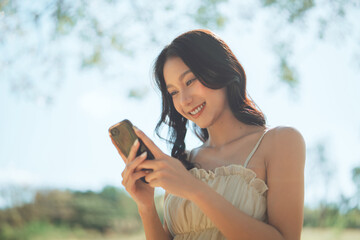 Smiling beautiful Asian woman using smartphone in public park, happy young lady checking social media or messaging on mobile phone outdoors, 5G technology and digital lifestyle concept.