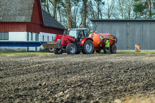 Falkenberg, Sweden, march 19-2025: A red tractor is seen maneuvering a slurry tank across an open farm field, preparing the soil for planting in early spring, surrounded by trees and buildings