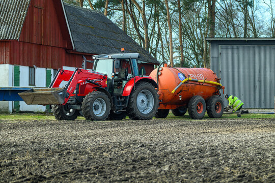 Falkenberg, Sweden, march 19-2025: A red tractor is seen maneuvering a slurry tank across an open farm field, preparing the soil for planting in early spring, surrounded by trees and buildings