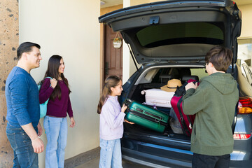 Family with teenage children packing luggage into car trunk, preparing for a summer road trip vacation