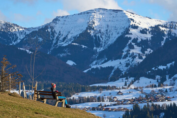 Senior woman hiking in winter near Oberstaufen, enjoying warm afternoon sunlight and a panoramic snowy mountain landscape in the Allg&auml;u region of Bavaria, Germany.