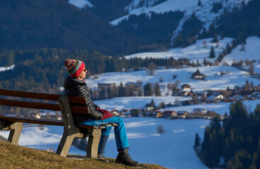 Senior woman resting on a bench during a winter hike near Oberstaufen, enjoying warm afternoon sunlight and a  snowy mountain landscape below Mount Hochgrat in the Allg&auml;u region of Bavaria, Germany.