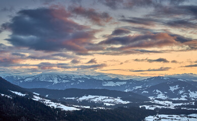 Alpine winter landscape in Bregenzer Wald at sunset with snow-covered mountains glowing in warm evening light under a dramatic sky.