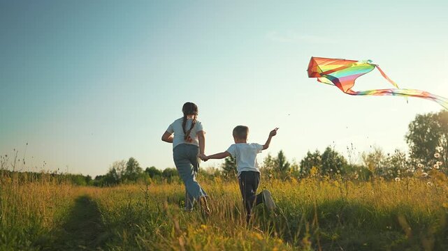 Girl and boy run holding hands flying rainbow kite in meadow at sunset outdoors. Sister with brother plays using colorful kite. Siblings run in field. Girl and boy fly kite. Children in meadow play.