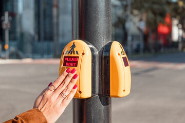 Woman's hand with red nails pressing a yellow pedestrian crosswalk button showing PLEASE WAIT in an urban setting