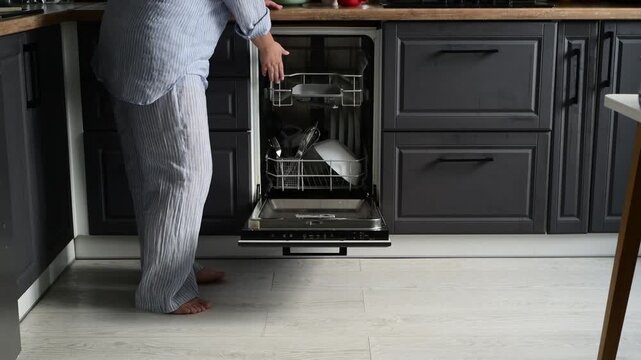 Woman in pajamas unloads a dishwasher and takes out clean kitchenware including pot, plates, cups, and glass bowls. Rear view with cropped frame showing hands, shoulders, and waist only.