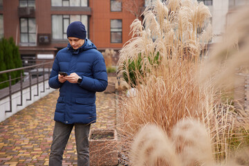 Man in winter coat and beanie texting on phone outdoors near ornamental grass