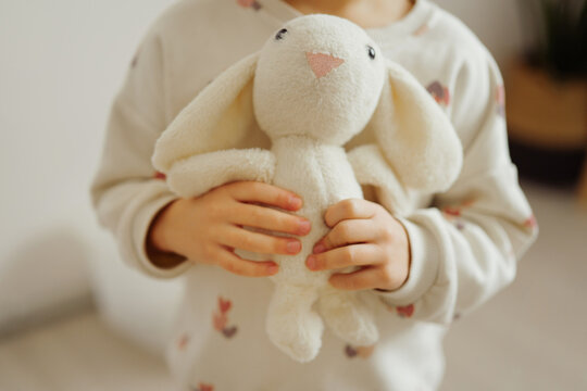 A child is holding a soft bunny toy in their hands while standing in a light room. The child wears a comfortable sweatshirt