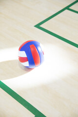 Single volleyball resting on polished hardwood gym court, showing red white blue panels and shadow