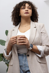 African American woman late twenties holding tan ceramic mug wearing beige blazer against pale wall