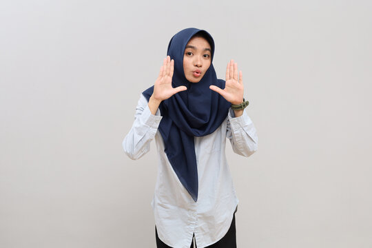 A shocked and dumbfounded young Asian woman in hijab with her palms next to her face, looking at the camera with a funny expression, reacting to something very surprising.