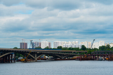 Blue subway train crossing a bridge over the river against a backdrop of modern residential buildings and construction cranes under a cloudy sky
