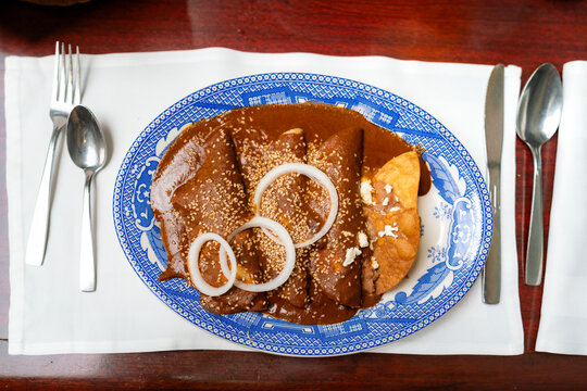 Classic enchiladas de mole served at Sanborns de los Azulejos in Mexico City, topped with sesame seeds and onion rings, accompanied by refried beans and tortilla on a traditional blue-patterned plate.