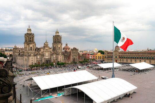Wide-angle view of the Z&oacute;calo in Mexico City with the iconic Metropolitan Cathedral, National Palace and waving Mexican flag under cloudy skies, showcasing colonial architecture and cultural grandeur
