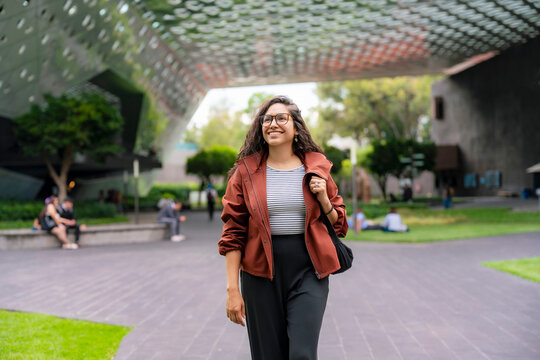 Young smiling Latina woman walks with energy and confidence outside the Cineteca Nacional in Coyoacan, Mexico City; wearing rust jacket, striped shirt, black pants and glasses on a bright day