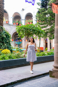 Latina woman walks calmly and smiling through a colonial courtyard in Centro de Coyoacan, Mexico City. Surrounded by plants, trees, papel picado and a fountain. She wears a white dress and sneakers