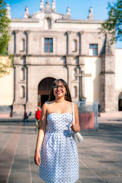 Young Latina woman walks confidently with a big smile in Centro de Coyoacan, Mexico City. Surrounded by flowerbeds and trees, wearing sunglasses and a strapless white dress with a blue print