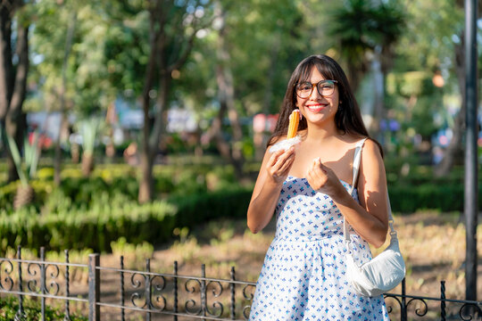 Happy young Latina woman smiles, she enjoys a churro relleno in a sunny park at Centro de Coyoacan, Mexico City. Wearing a white printed dress and glasses, surrounded by trees, plants and iron fences