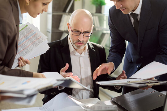 Stressed manager with colleagues facing paperwork overload pressure. Documents pile up as the team demands decisions, fueling frustration and deadline stress. Strong concept of workplace burnout.
