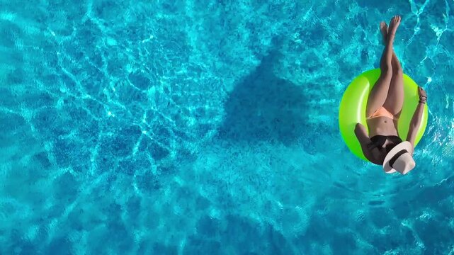 Aerial view of young woman in hat relaxing in swimming pool, beautiful girl in bikini floats on inflatable ring donut and has fun in water, tropical vacation on holiday resort, top drone view