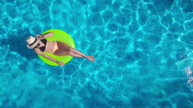 Aerial view of young woman in hat relaxing in swimming pool, beautiful girl in bikini floats on inflatable ring donut and has fun in water, tropical vacation on holiday resort, top drone view