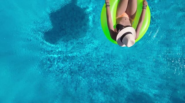 Aerial view of young woman in hat relaxing in swimming pool, beautiful girl in bikini floats on inflatable ring donut and has fun in water, tropical vacation on holiday resort, top drone view
