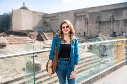 Young Latina woman smiles calmly next to the ancient ruins of Templo Mayor behind the Cathedral in Mexico City, wearing blue cardigan, jeans with bright sunlight and cultural landmarks surrounding her