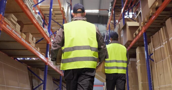 Warehouse workers carrying boxes between shelves. Men in vests move along racks, handling packages for delivery, shipping and inventory. Concept shows efficient warehouse order fulfillment.