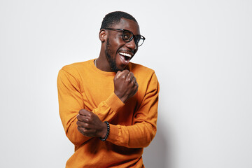 African American man with dark skin wearing glasses and orange sweater smiling and celebrating with clenched fists on white background studio portrait.