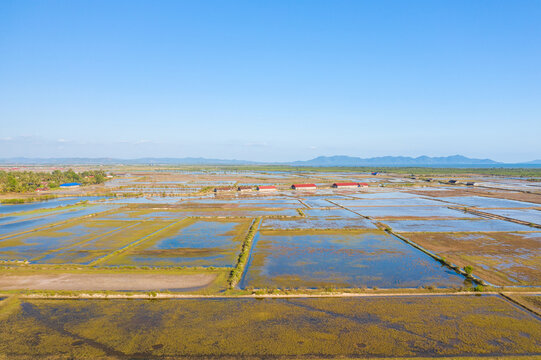 Expansive salt evaporation ponds stretch across the landscape near Kampot, Cambodia, with reflective water surfaces and geometric patterns visible under a clear sky. Distant red-roofed sheds and a