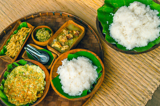 Traditional Thai food is served in wooden bowls with round and square shapes on a bamboo tray with spoons. There is plain rice, omelet, curry, and chili paste on a woven mat with soft sunlight.