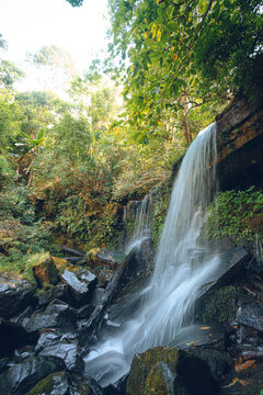 A waterfall flows down from a cliff in the forest. The water is strong in the rainy season. There are black rocks, trees, and moss all around. Idea for a natural background in soft tones.