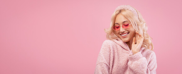 Fototapeta premium A young woman enjoys a moment while wearing a fluffy pink hoodie and pink sunglasses. She poses playfully in front of a pink background and has long blonde hair.