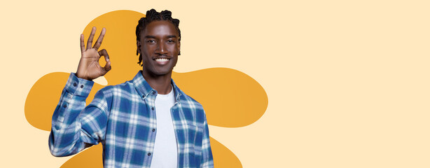 A young man stands and smiles while making an okay hand gesture. He wears a plaid shirt and is in front of a plain yellow background. His expression shows confidence and positivity. © Prostock-studio