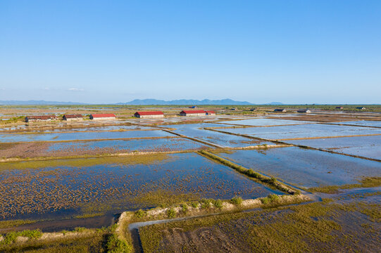 Rows of rustic salt storage sheds with red roofs line the edge of expansive flooded salt evaporation ponds near Kampot, Cambodia. The landscape features geometric water patterns, open sky, and distant