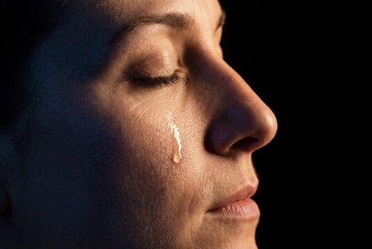 Close up portrait of a middle aged woman with a single tear rolling down her cheek, expressive eyes closed in grief or sadness, dramatic side lighting on dark background