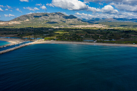 Aerial view of deep blue sea meeting a sandy beach and breakwater, with lush green countryside and rugged mountains under scattered clouds near Orosei, Sardinia.