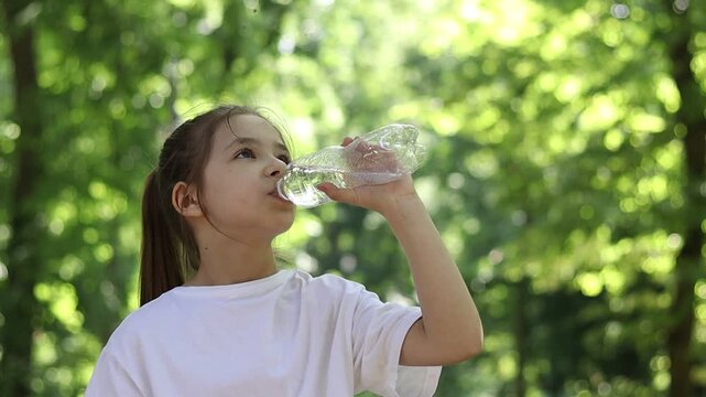 A girl stays hydrated by drinking water from a bottle in a green summer park.