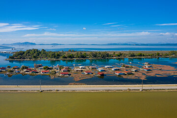 Fototapeta premium Colorful stilt houses line the edge of Tourlida lagoon in western Greece, surrounded by calm blue water and lush greenery under a clear sky. The tranquil wetland landscape features reflections, small