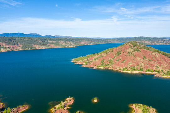 Aerial shot of a vibrant red hill topped with green vegetation jutting into the deep blue waters of Lac de Salagou, surrounded by rugged countryside and distant mountains under a bright sky.
