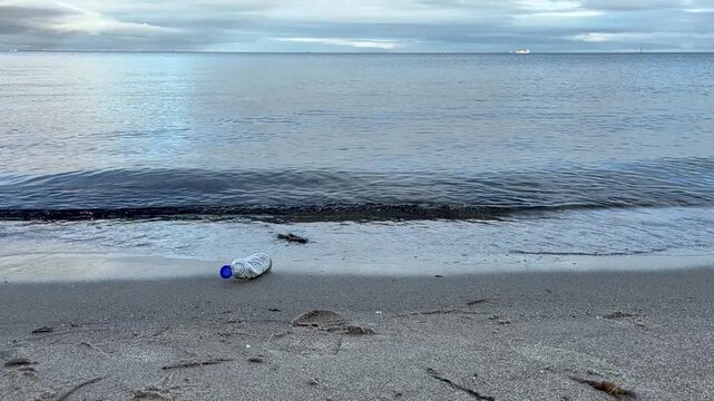 Young woman picks up plastic bottle at shoreline &ndash; environmental cleanup on beach
