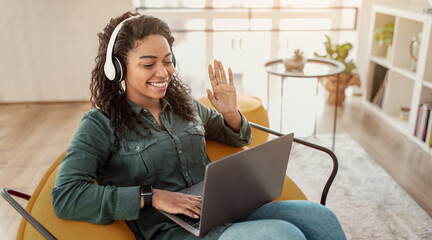 A woman sits in a chair in her living room. She wears headphones and has a laptop in front of her. She is smiling and waving while on a video call. Sunlight enters through the window. © Prostock-studio